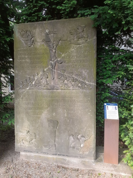 Grabplatte für Johann Brüggmann von 1720 mit dem Bild eine Kruzifix, das von den männlichen und weiblichen Familienmitgliedern angebetet wird; darunter als Berufszeichen ein Stiefel, in den Ecken Evangelistensymbole.